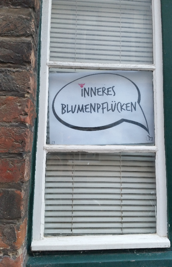 ein altes Fenster, drin hängt eine
auf Papier ausgeschnittene Sprechblase mit dem Text 'inneres Blumenpflücken'
